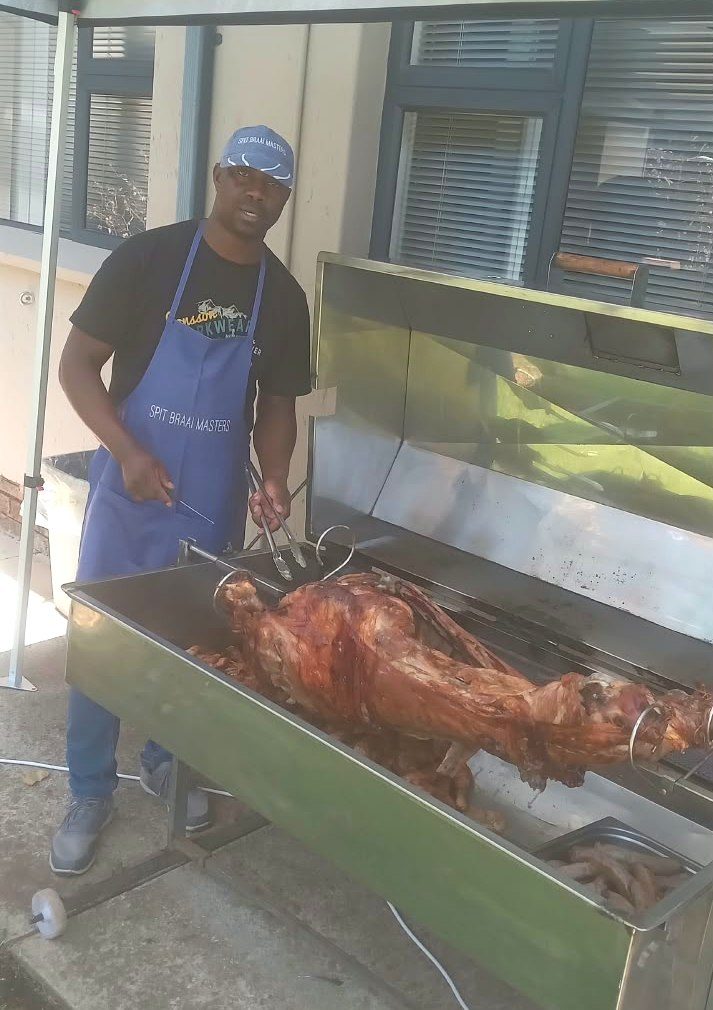 Chef Joseph in a Spit Braai Masters cap and apron, turning a whole lamb on the spit at a function